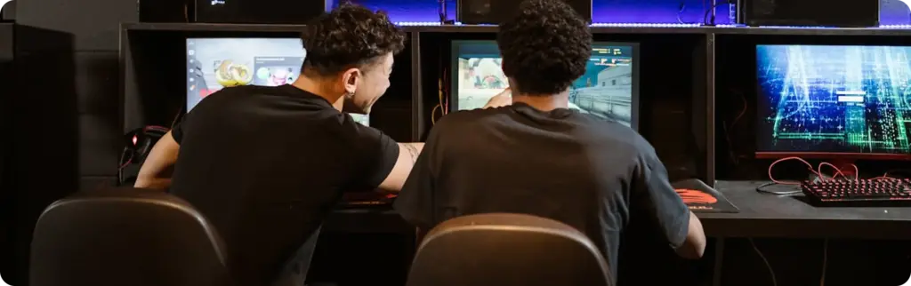 Twee gamers die op desktopcomputers spelen in een LAN-gamingcentrum, zij aan zij zittend met meerdere beeldschermen in een sfeervol verlichte esports-omgeving.