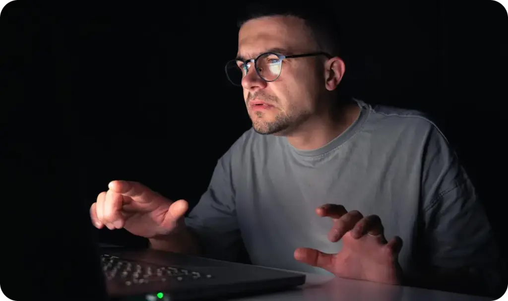 Een man met een bril die ’s avonds achter een laptop zit, intens geconcentreerd in een donkere kamer, verlicht door het schermlicht terwijl hij aan het gamen is of aan het werk is.